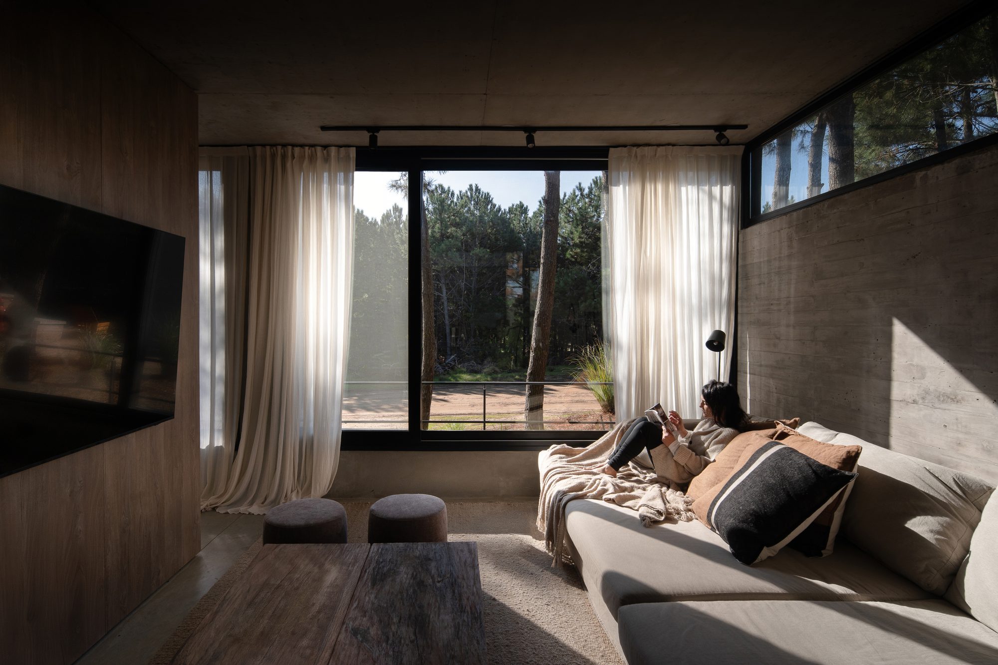 Living con sofá y cortinas, ventanal corrido al pinar bajo losa de hormigón — CORNER Arquitectura