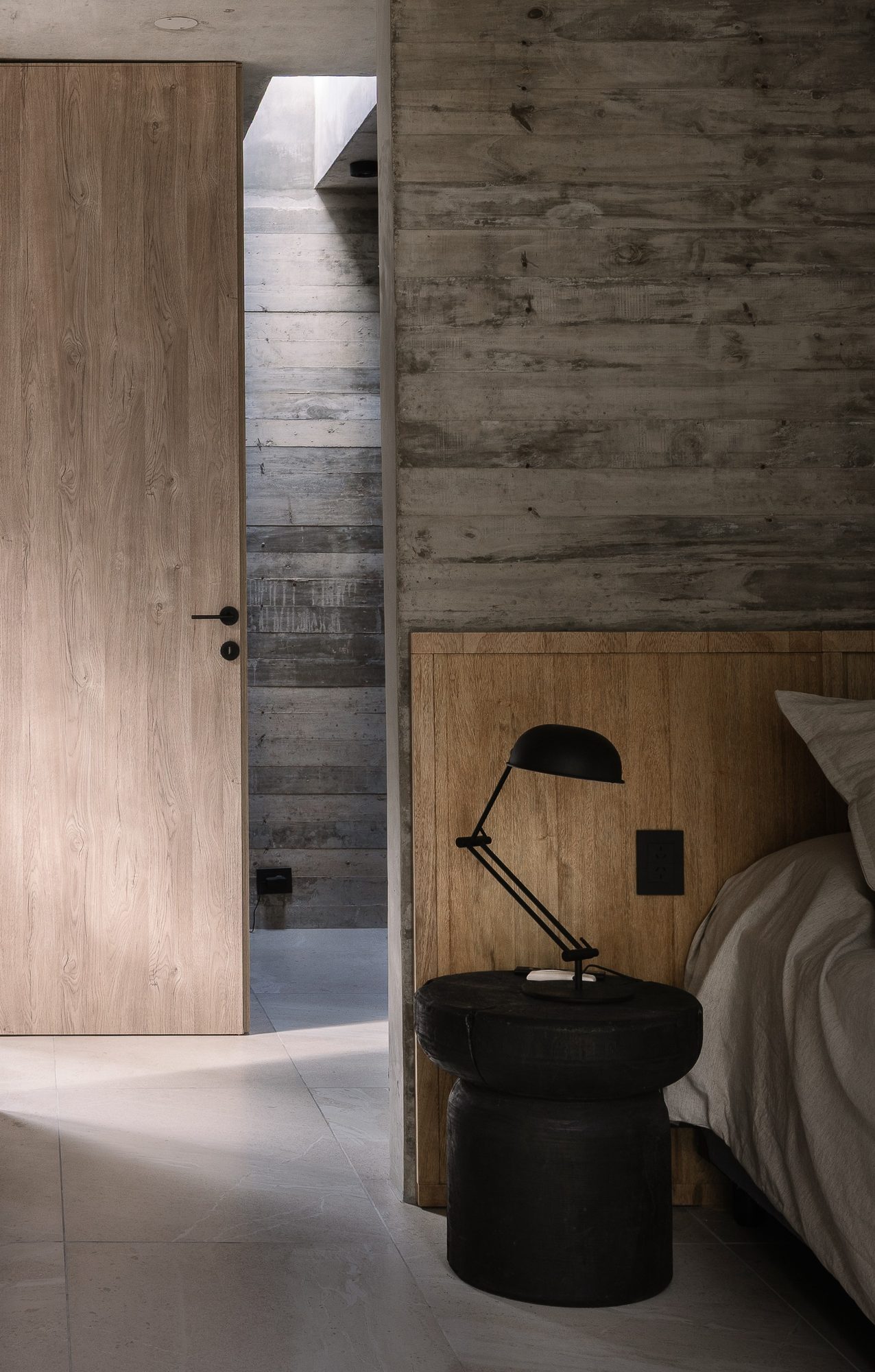 Dormitorio con mesa de luz negra y velador, entre muros de hormigón y madera — CORNER Arquitectura