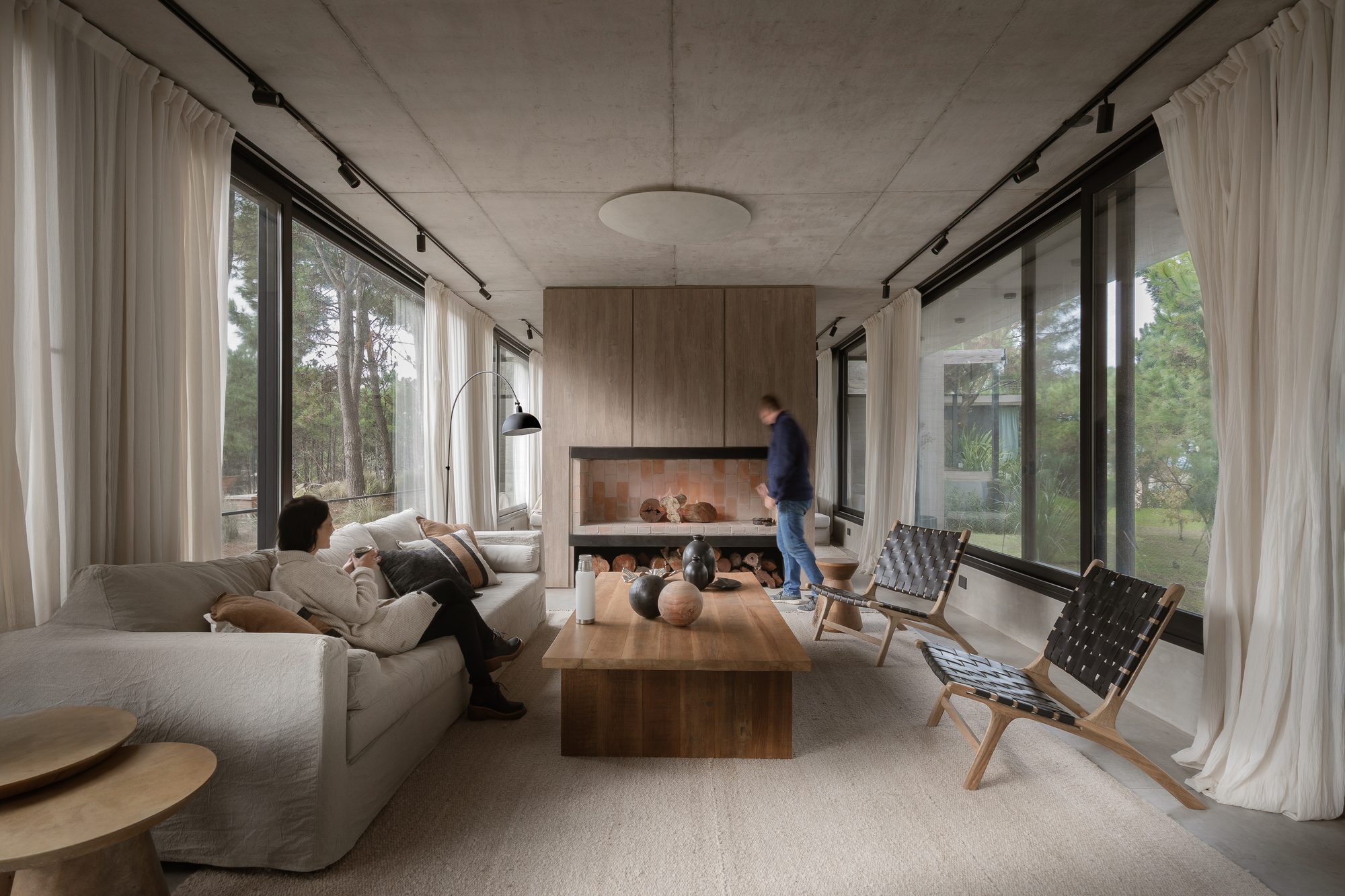Living con hogar a leña, cortinas blancas y sillones frente al ventanal al bosque — CORNER Arquitectura