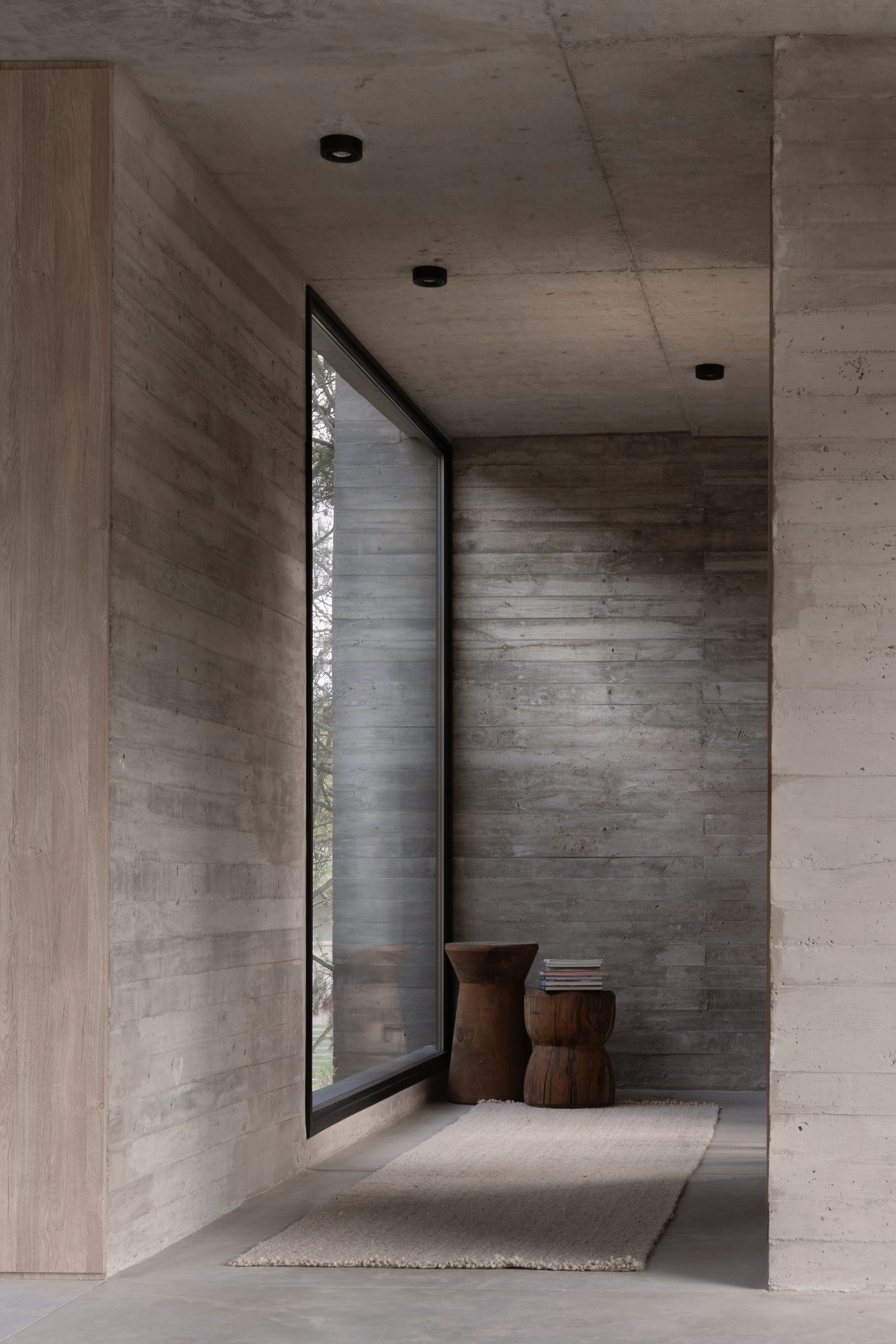 Pasillo de hormigón visto con ventanal vertical al fondo y banquetas de madera — CORNER Arquitectura