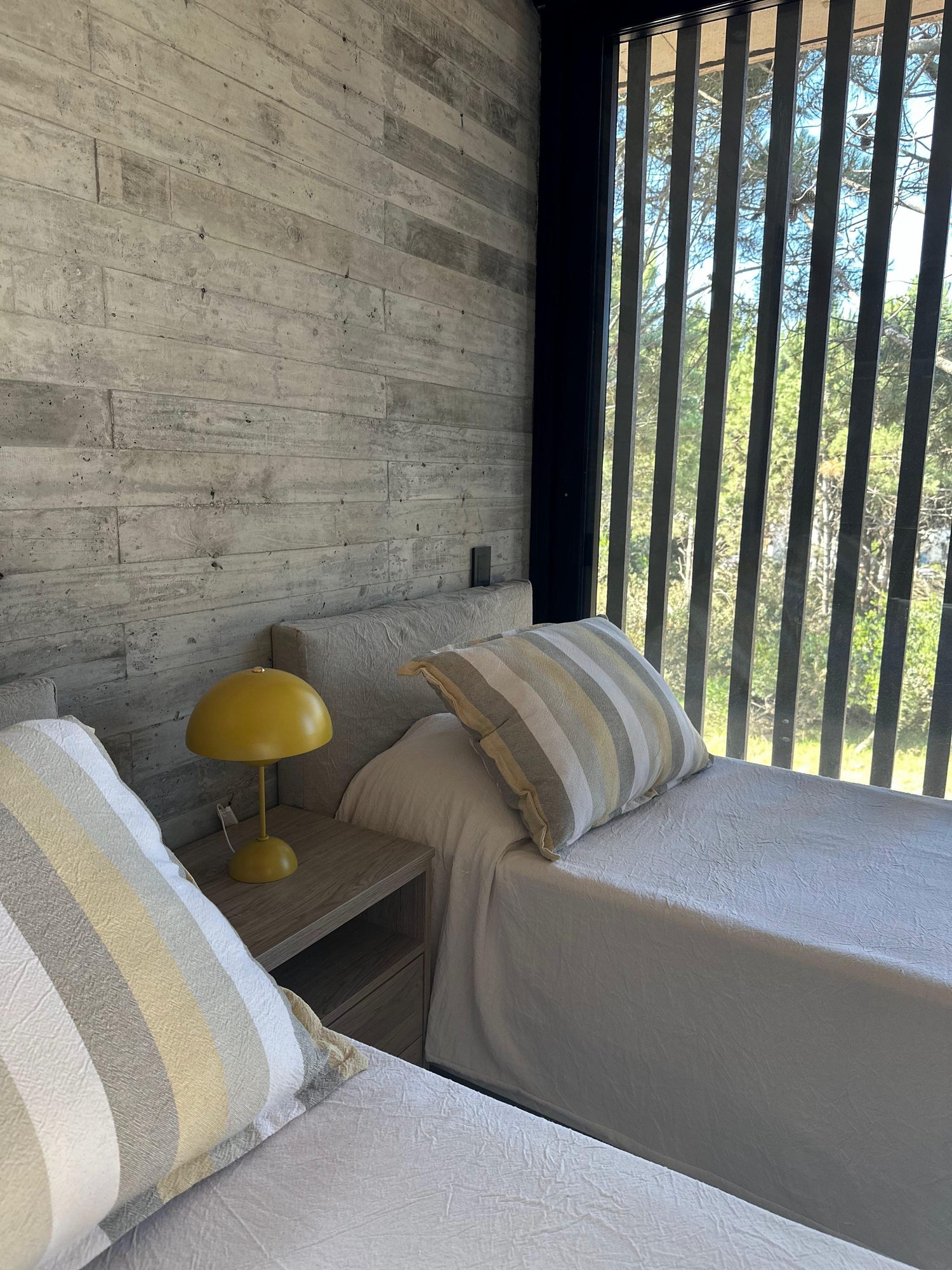 Dormitorio con velador amarillo y ropa de cama a rayas, junto a la celosía — CORNER Arquitectura