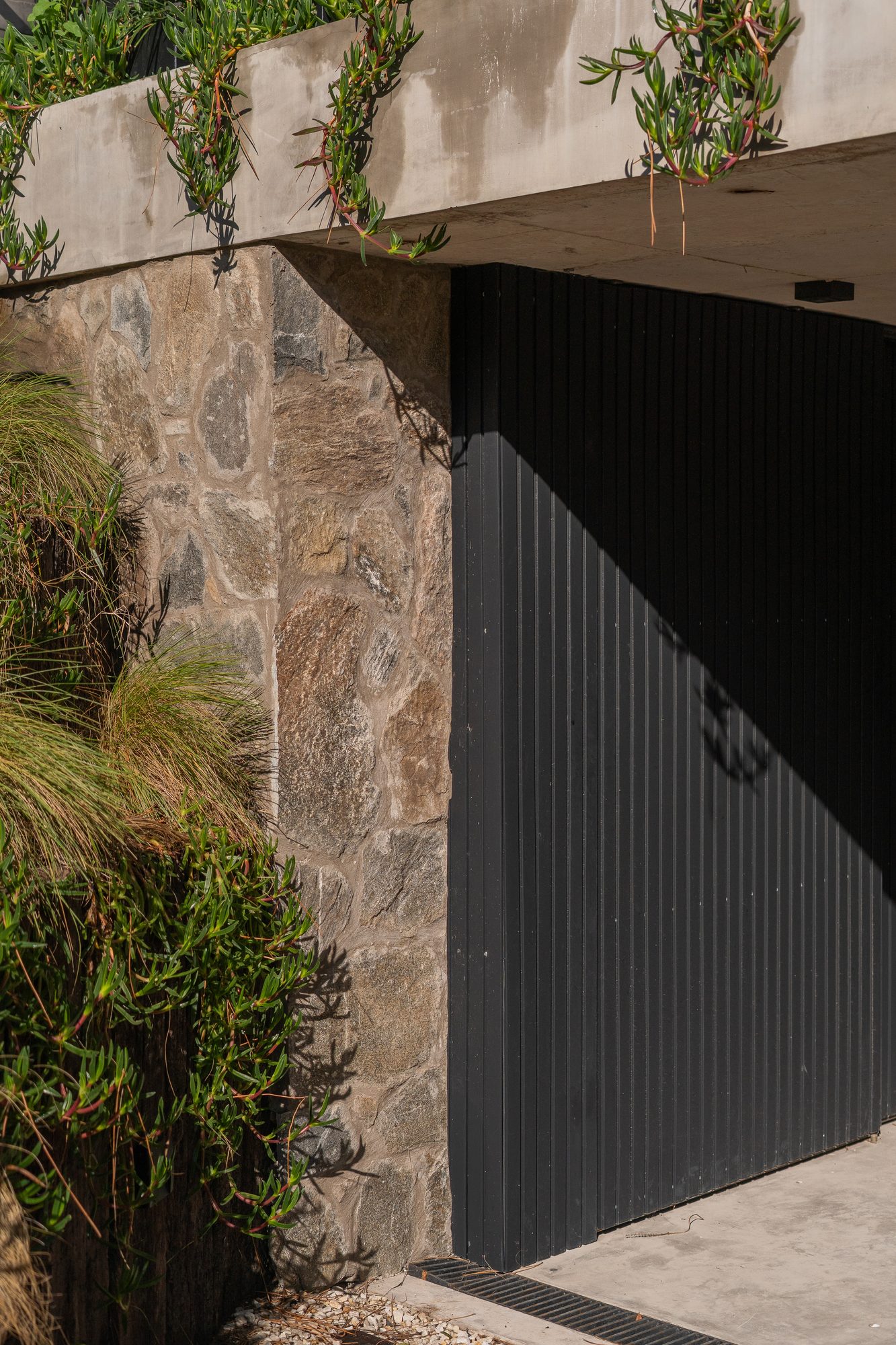 Acceso con portón de chapa plegada entre muro de piedra y vegetación — CORNER Arquitectura