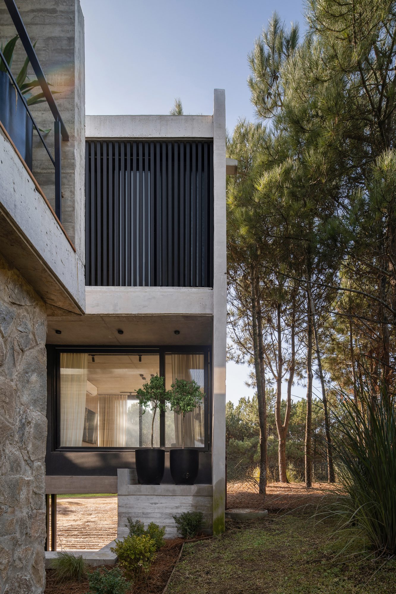Fachada de dos niveles con balcón y parasol, rodeada de pinos — CORNER Arquitectura