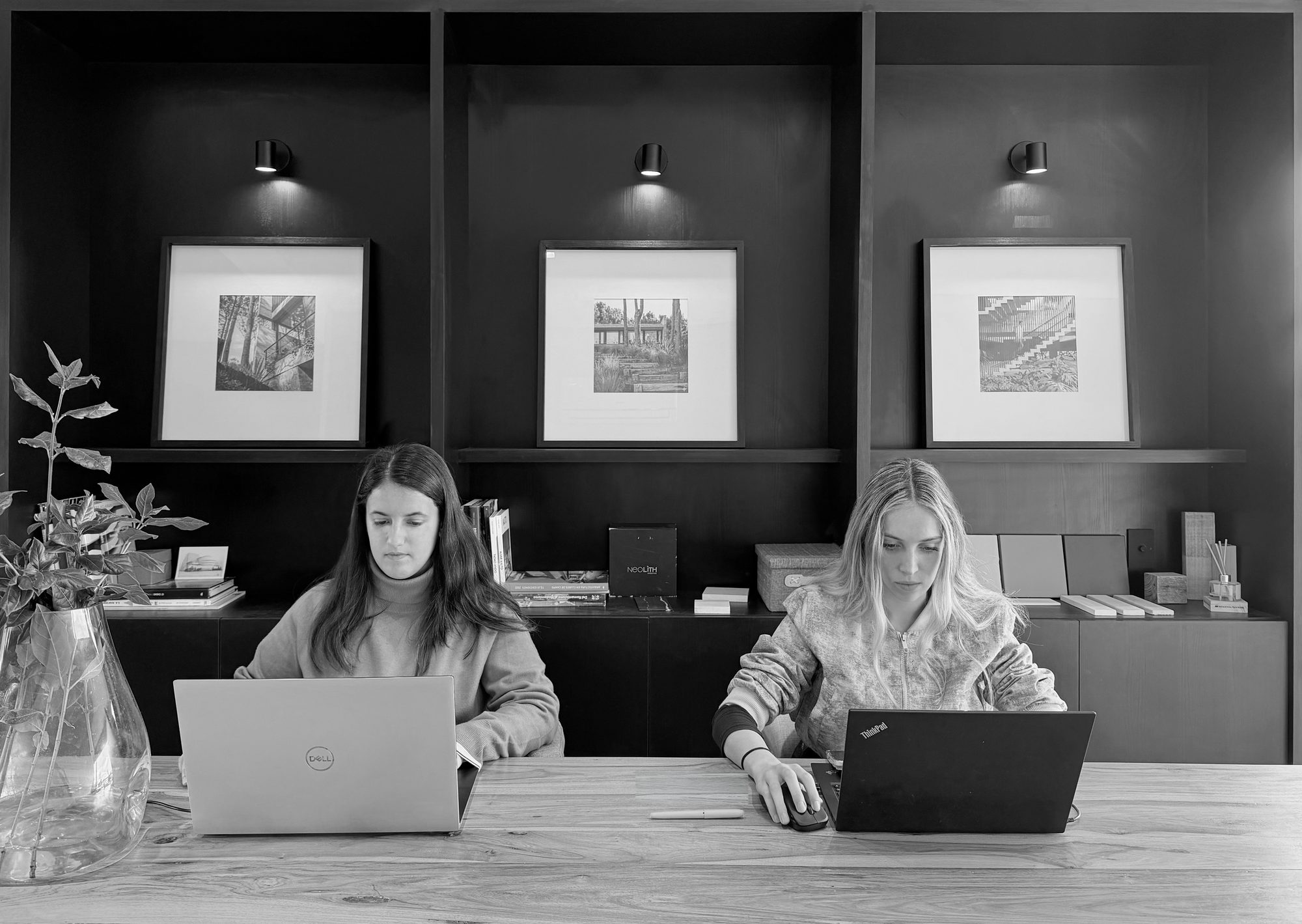 Dos integrantes del equipo trabajando en el estudio, frente a la biblioteca con láminas enmarcadas — CORNER Arquitectura